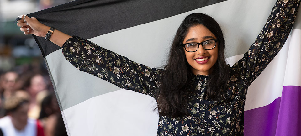 A person with brown skin, long dark hair, and a black floral shirt is holding an ace flag behind them with their outstretched arms.