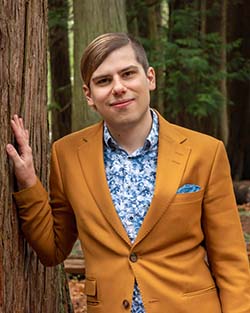 Headshot of Basil, a white nonbinary person with an undercut, wearing a mustard blazer and patterned blue shirt.