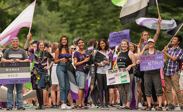 A diverse group of people are smiling and holding a variety of ace and aro flags and signs.