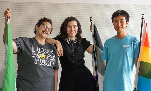 Three people stand side by side in front of a gray wall. Each holds a different pride flag.