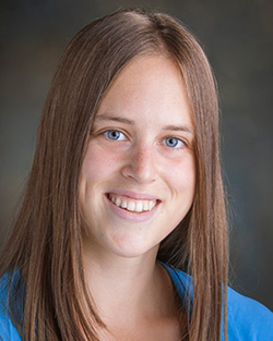 Headshot of Justine, a white genderfluid person with shoulder length brown hair wearing a blue shirt.